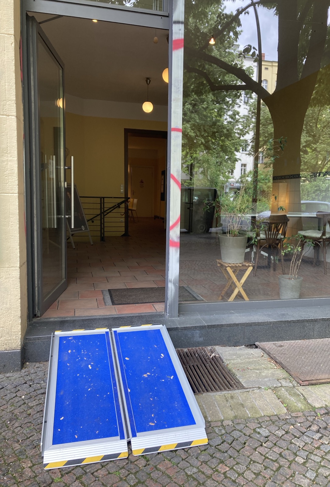 Eingang IQZ Yorckstraße Verglaste Ladenzeile mit geöffneter Tür und angelegter Rampe. Im Hintergrund sieht eins die Projekträume IQZ. In der Glasfront spiegeln sich Bäume der Yorckstraße.